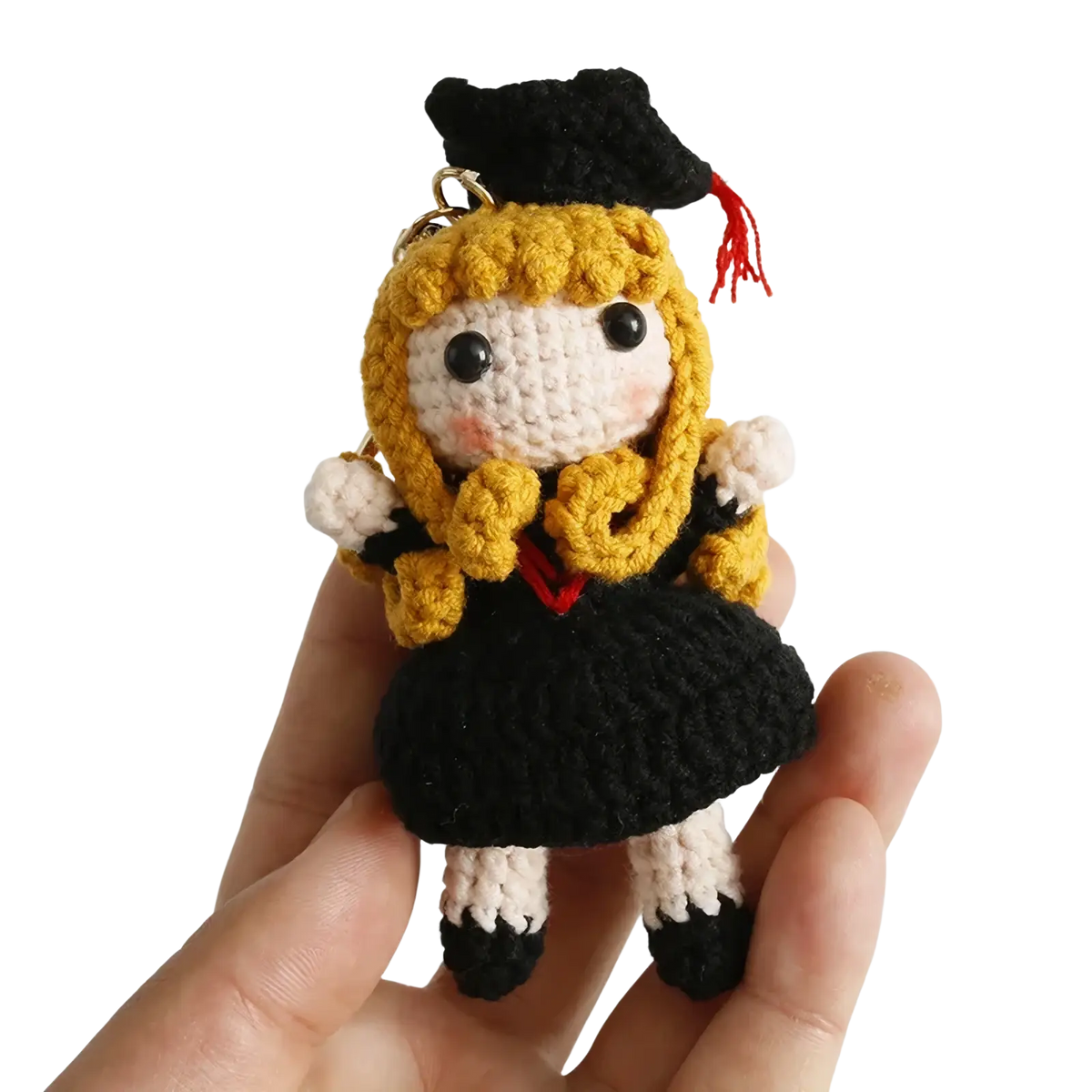 A crocheted graduation doll keychain featuring a blonde-haired figure in black gown and cap with red tassel and white accents