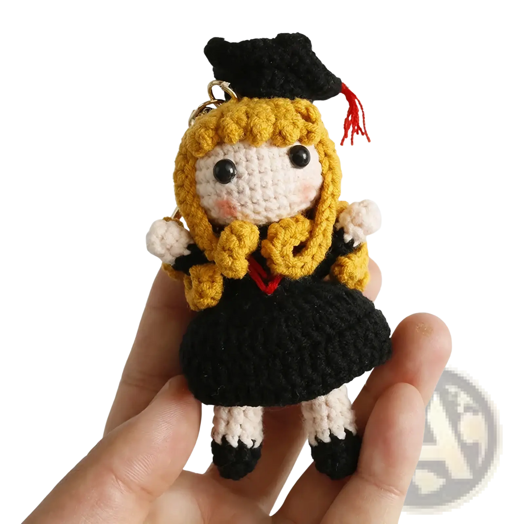 A crocheted graduation doll keychain featuring a blonde-haired figure in black gown and cap with red tassel and white accents