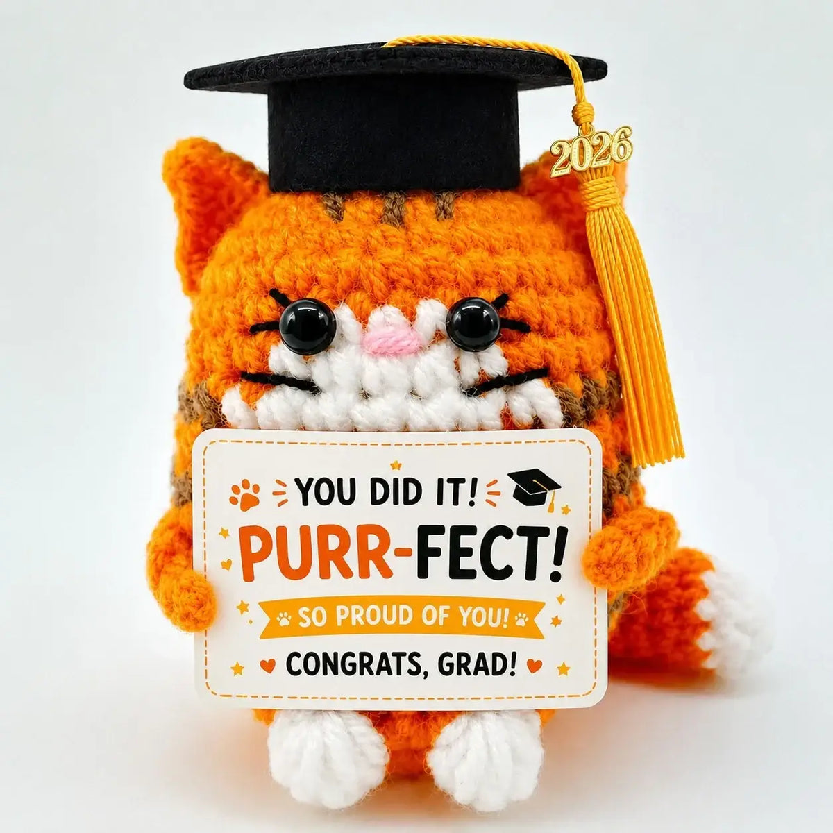 Orange crocheted cat keepsake wearing black graduation cap with yellow tassel holding white congratulatory sign