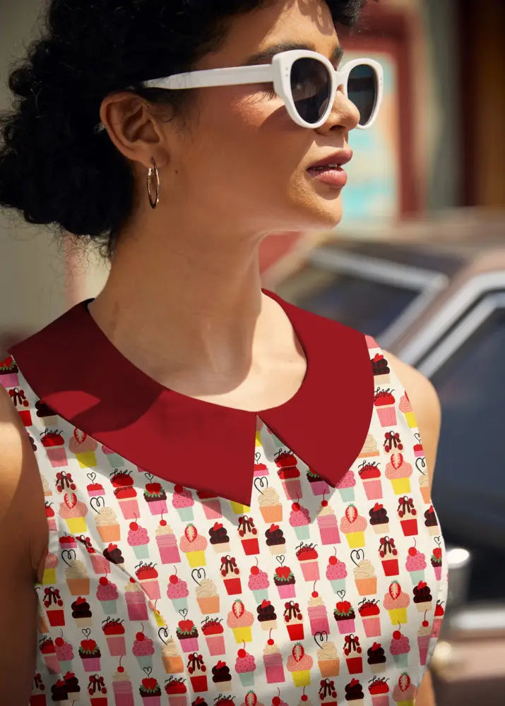 A woman wears a retro cupcake print sleeveless cotton dress with pockets featuring a white base and red collar