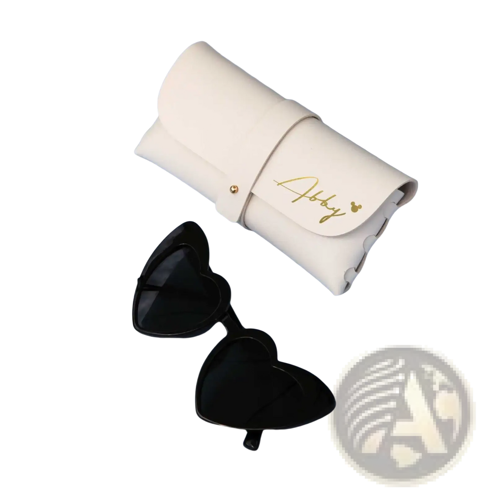 Black heart-shaped sunglasses with dark lenses next to a cream leather glasses case with gold script and button