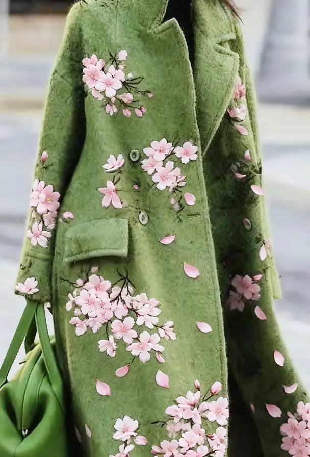 Ein weich schimmernder olivgrüner Wollmantel für Damen mit zarter rosa Kirschblütenstickerei und verstreuten Blütenblättern.