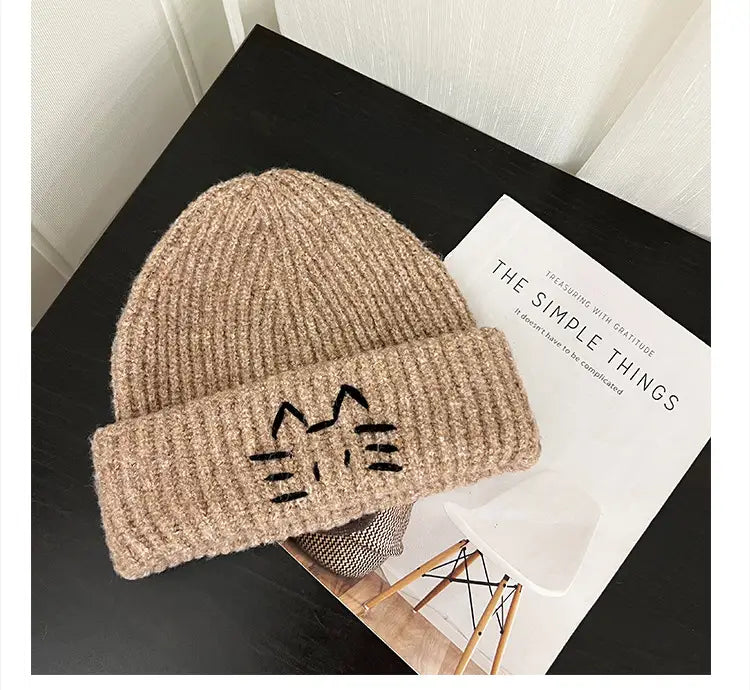 A cozy beige knitted beanie hat featuring a cute black embroidered cat face on the folded brim.