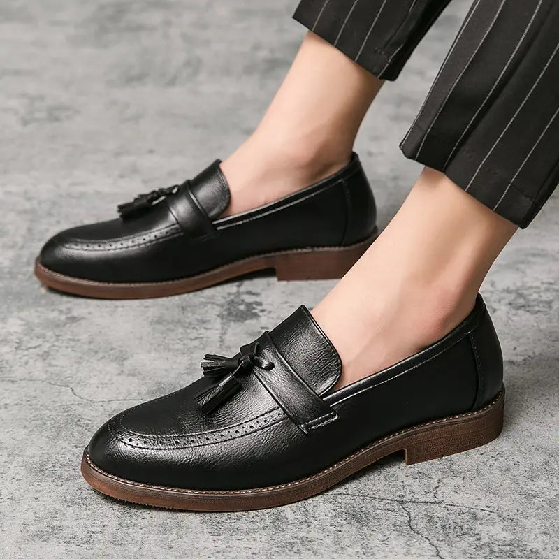 Mocasines de cuero tipo brogue con borlas para hombre en negro con suelas de goma marrones y acentos brogue detallados.