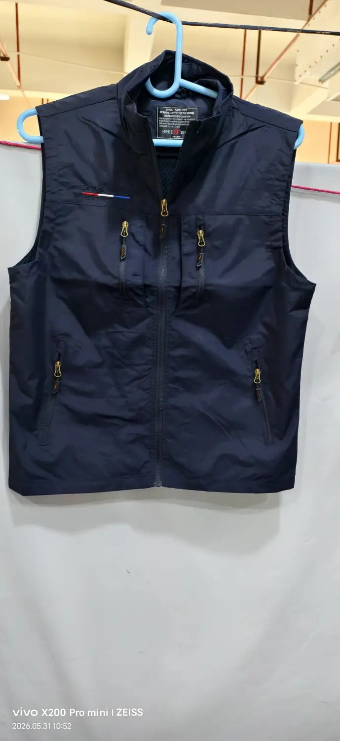 Chaleco utilitario con capucha y múltiples bolsillos para hombre en azul marino oscuro sin mangas, con cremalleras tono dorado y un pequeño logotipo rojo, blanco y azul en el lado izquierdo del pecho.