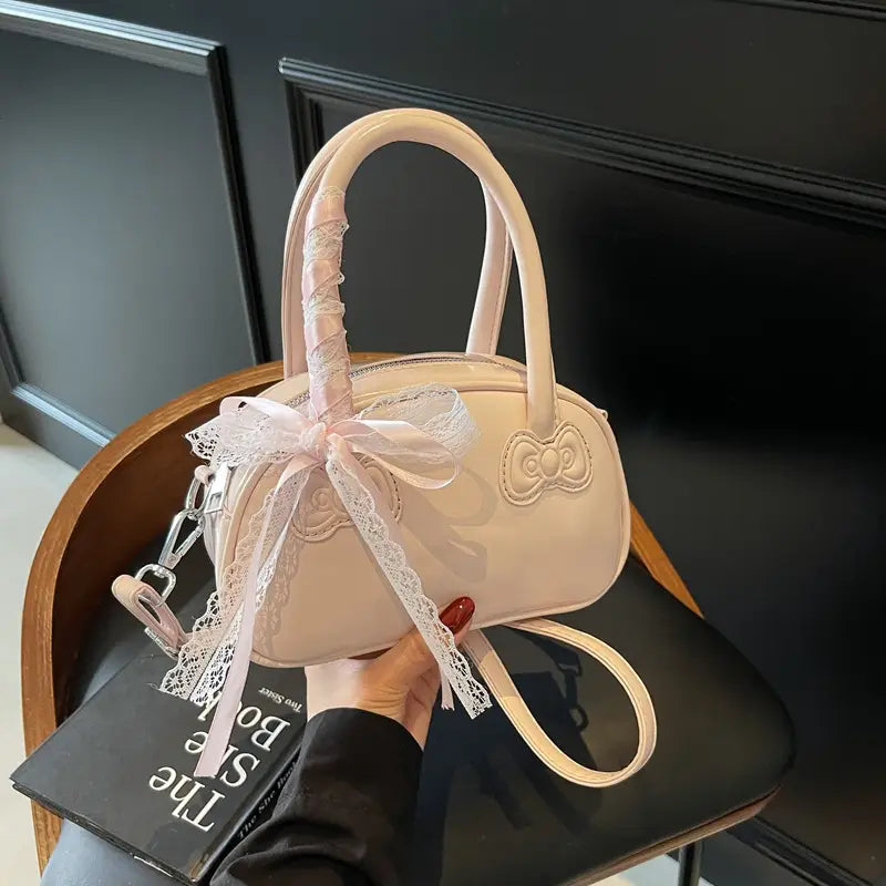 A blush pink glossy crossbody bag featuring a bow-shaped handle, lace trim, and embroidered bow detail from the Hello Kitty collection.
