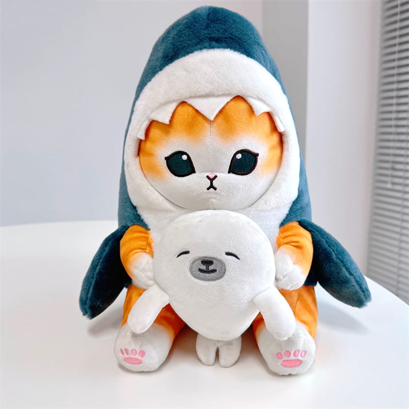 オレンジと白の毛並みの猫がティール色のサメのフーディーを着用し、小さな白いアザラシのぬいぐるみを抱えた「猫サメ」ぬいぐるみキーホルダー。日本の猫の人形（だるま）風のペンダントとしてデザインされています。