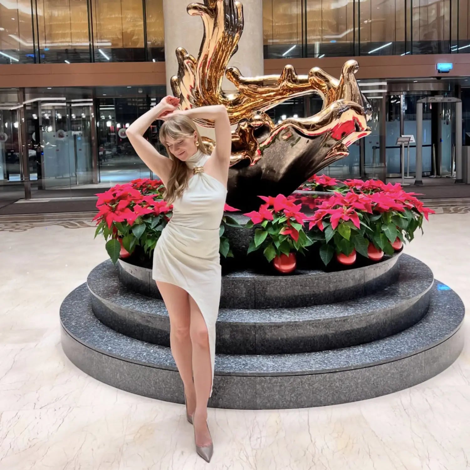 A woman wearing a sleek off-white gold ring halter stretch mini dress with a high slit stands beside a golden abstract sculpture surrounded by poinsettias.