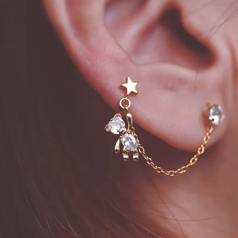 Boucles d’oreilles asymétriques en strass à finition dorée, avec ours et étoiles, comportant des pampilles, un pendentif étoile et un ours suspendu orné de cristaux transparents reliés par une chaîne à un cabochon en cristal.