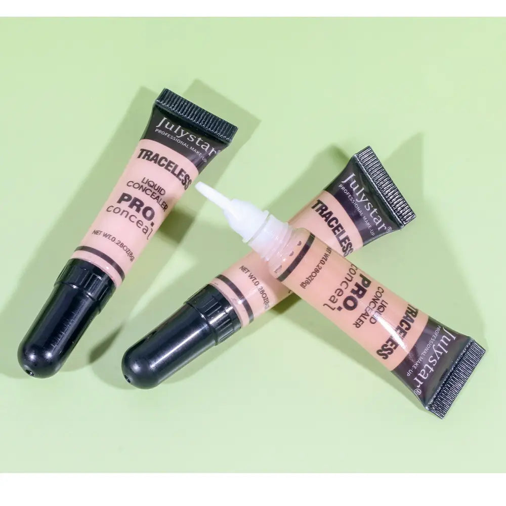 Two black-tipped peach-colored julystar traceless pro liquid tattoo concealer tubes with white applicator tips lie diagonally on a light green surface.