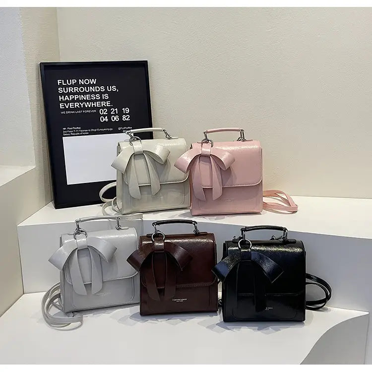 Vier kleine, glänzende Schleifen-Umhängetaschen in Weiß, Pink, Grau und Schwarz aus Kunstleder als modische Handtaschen.