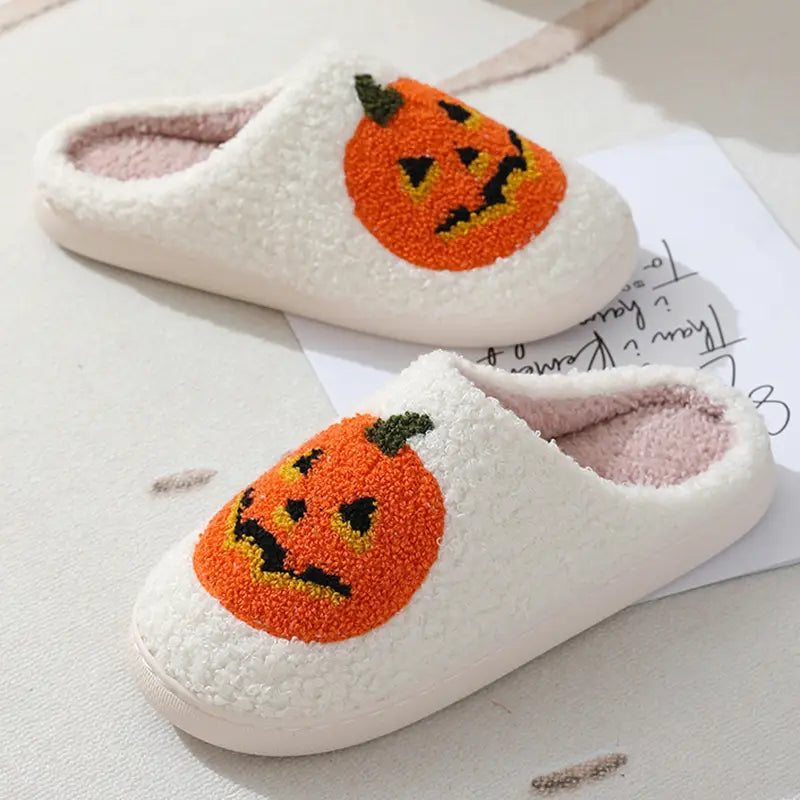 White plush slip-on slippers featuring a cheerful orange jack-o-lantern face with black and yellow embroidered details designed for Halloween.