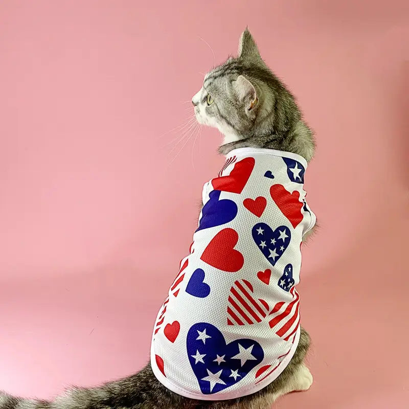 Un gato atigrado gris que lleva un chaleco de malla para perro con bandera estadounidense, camiseta para mascota en EE. UU., con material de malla blanca decorado con corazones en rojo, azul y con patrón de estrellas.
