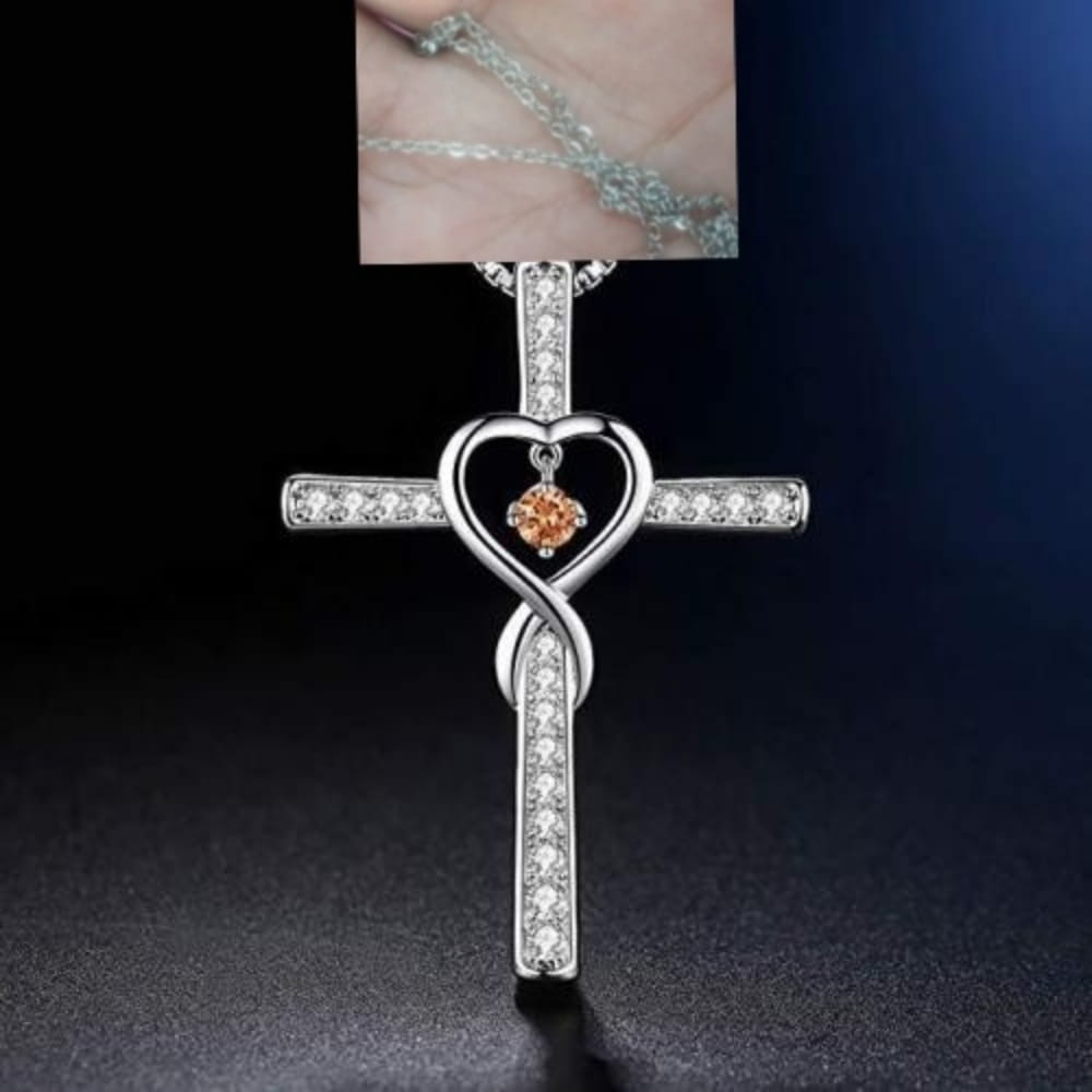 Silver cross pendant with a heart-shaped center holding an orange gemstone adorned with clear crystals and an intertwined design in the infinity love heart cross birthstone crystal necklace.