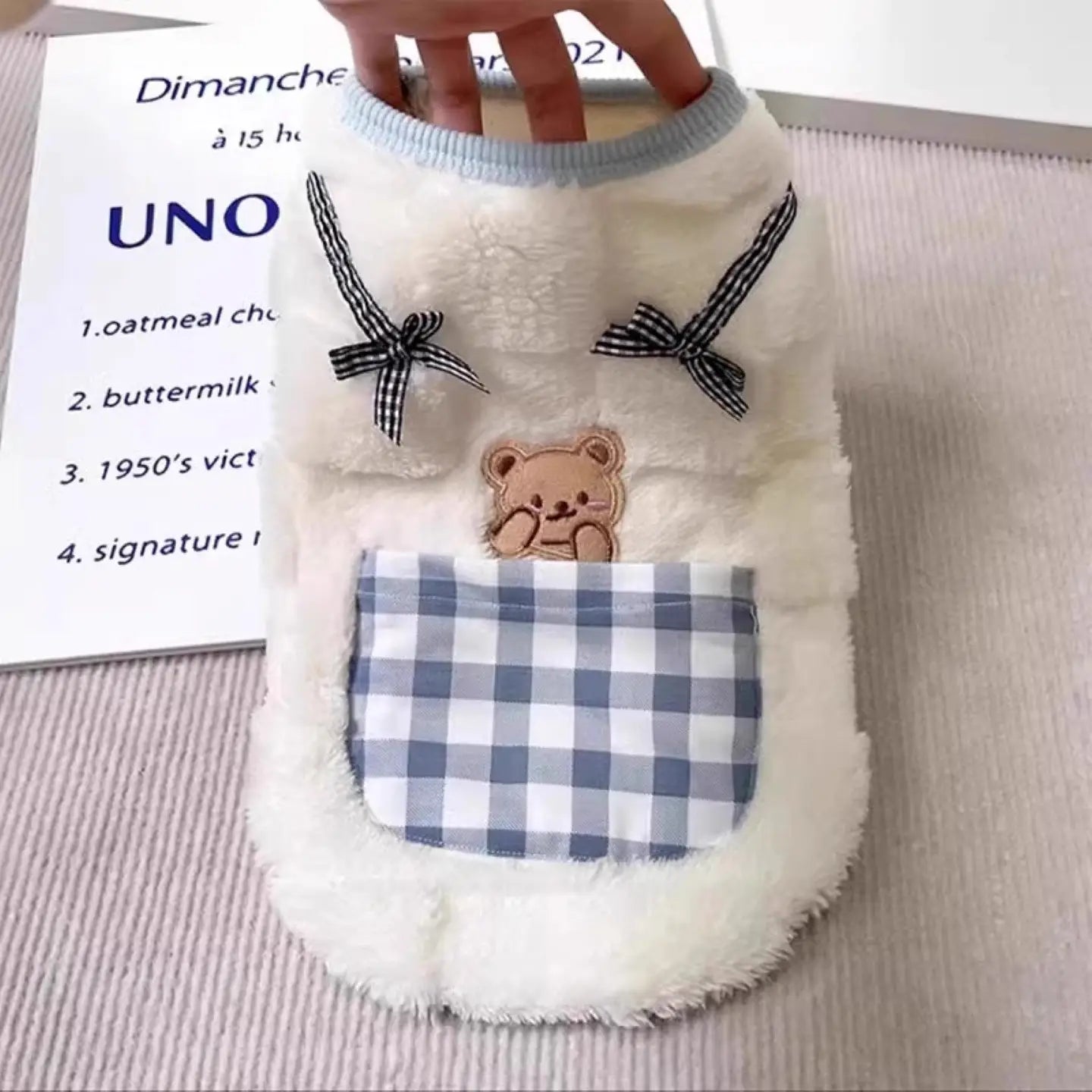 fluffy winter cat vest in white with black and white checkered bows featuring a teddy bear patch and a blue and white gingham pocket.