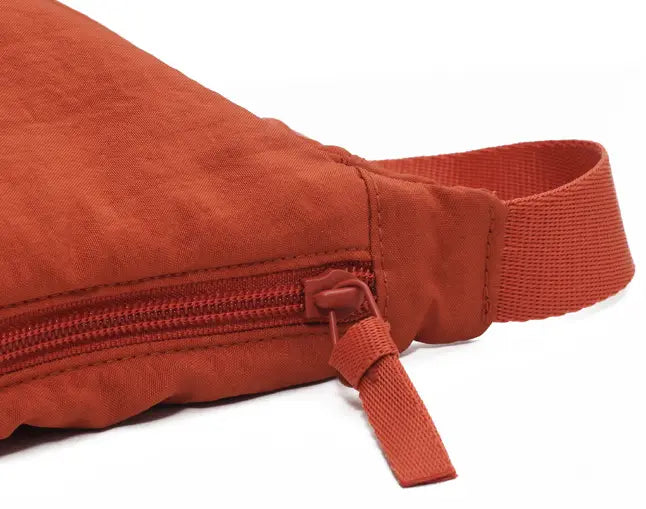 A rust-colored nylon dumpling messenger bag with a curved crossbody strap features a matching zipper and canvas fanny pack design.