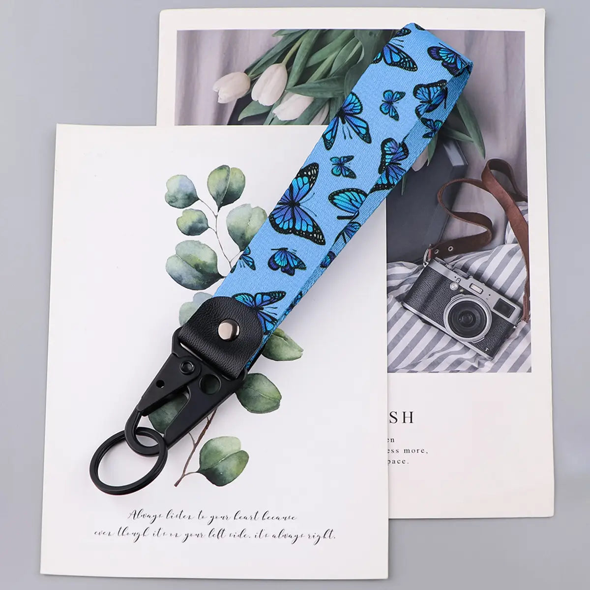 A light blue butterfly wrist strap keychain featuring a repeating pattern of black and teal butterflies attached to a black metal clip with a ring.