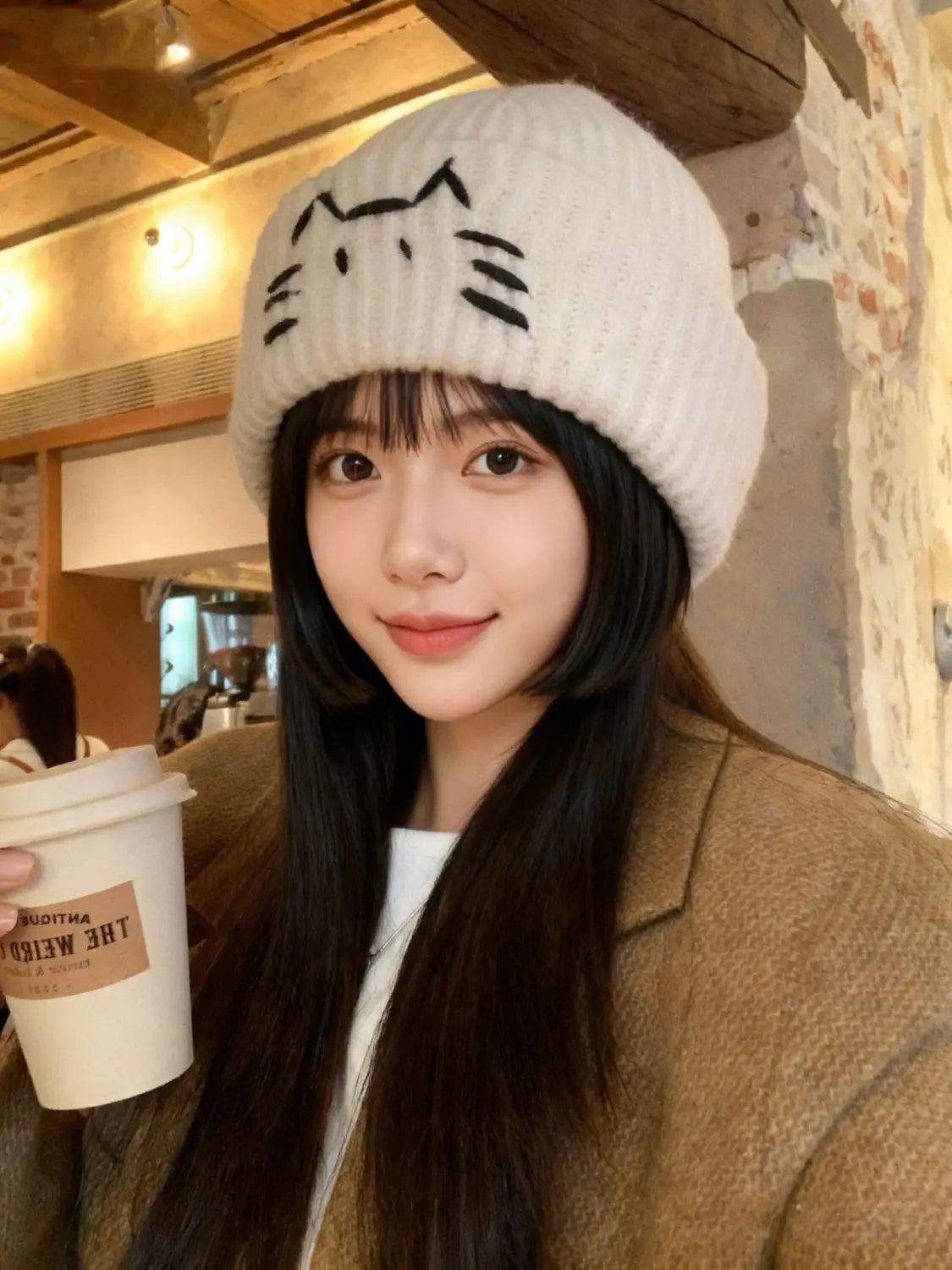 A soft off-white knitted beanie hat featuring a black embroidered cat face design.