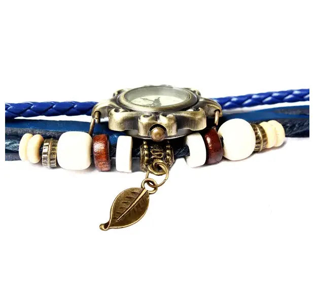 Antique brass-finished watch with a round face set on layered blue braided leather straps adorned with white brown and cream-colored beads and a leaf-shaped pendant in a Bohemian blue braided leather leaf charm wrap watch.