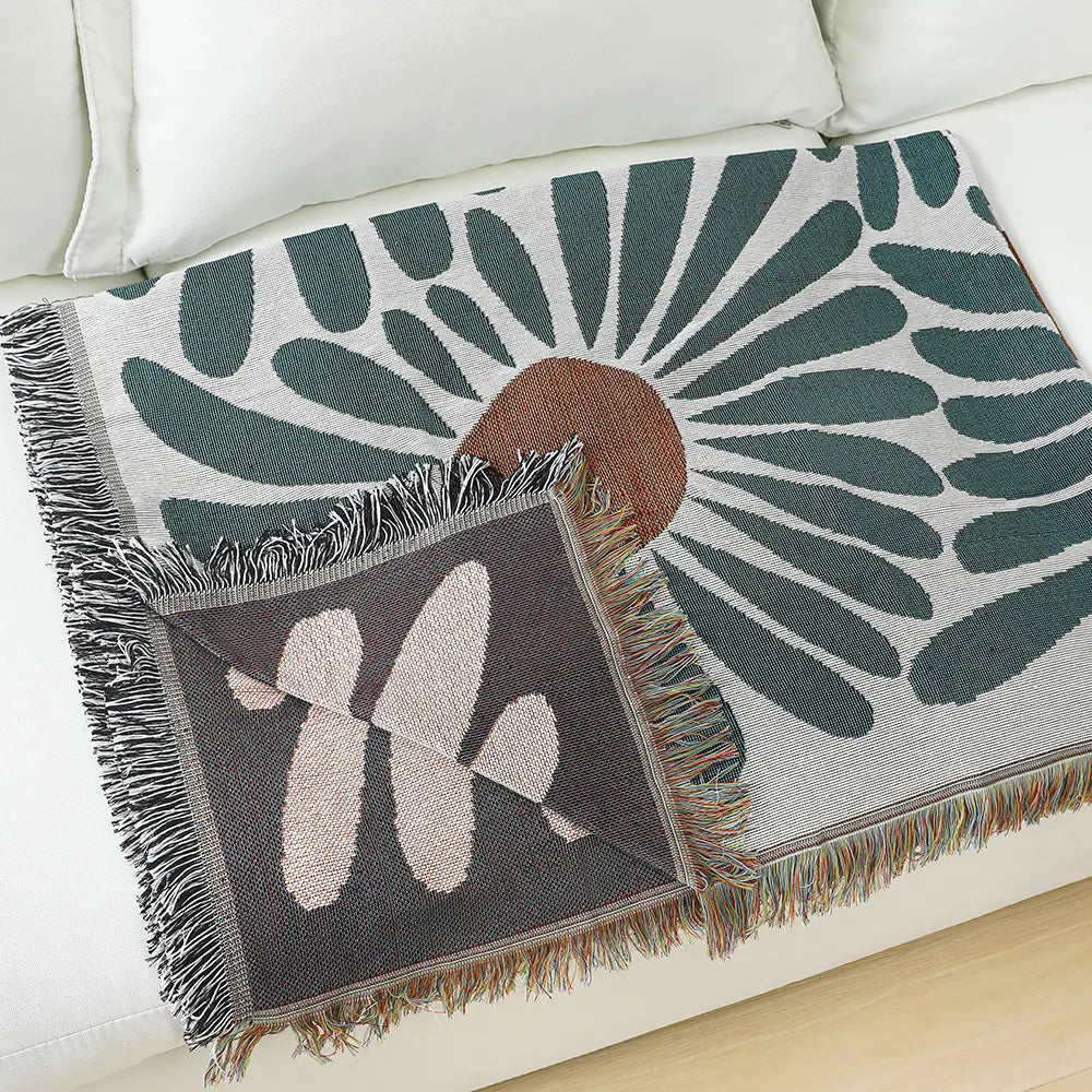 A woven cotton daisy and geometric floral throw blanket with a cozy textured design featuring dark green leaf-like patterns radiating from a central brown circle on a light gray background accented by fringed edges and a deep gray square patch with beige abstract shapes.