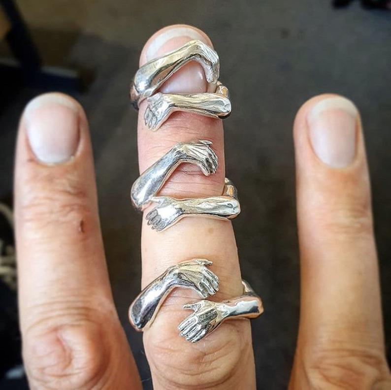 Adjustable silver ring featuring intricately designed intertwined hands with a polished metallic finish and a red gemstone.