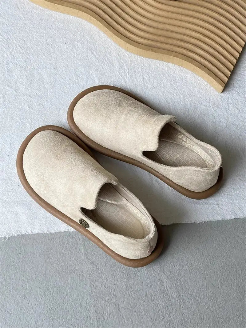 Beige Veloursleder-Slipper im Vintage-British-Stil mit niedrigem Absatz mit einer braunen Gummisohle und einer kleinen metallisch wirkenden Knopfdetailierung an der Seite.