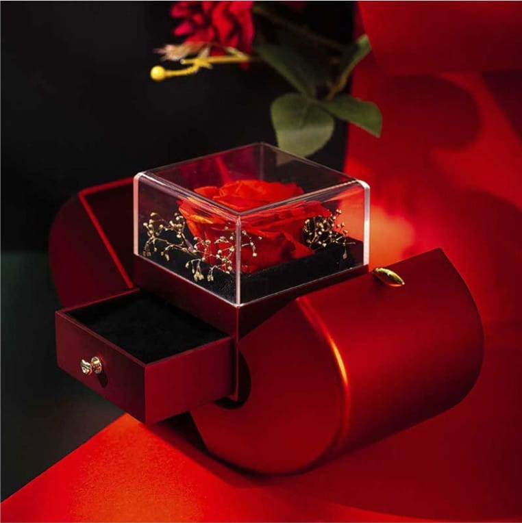 A vibrant red preserved rose inside a clear acrylic cube displayed on an elegant burgundy velvet eternal rose apple jewelry box with a hidden necklace drawer and gold-tone hardware.