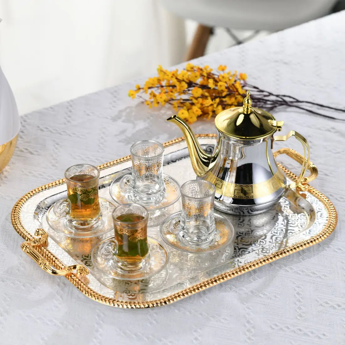 Luxury rectangle serving tray with gold handles featuring a gold and silver ornate tea set with intricate embossed patterns and clear glass cups with gold detailing.