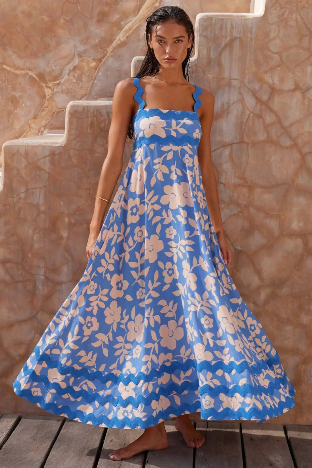 Un vestido maxi azul fluido con flores y patrones de hojas de color crema, que presenta escote cuadrado y tirantes de hombro festoneados, diseñado como un vestido maxi de playa en corte A con ribete ondulado.