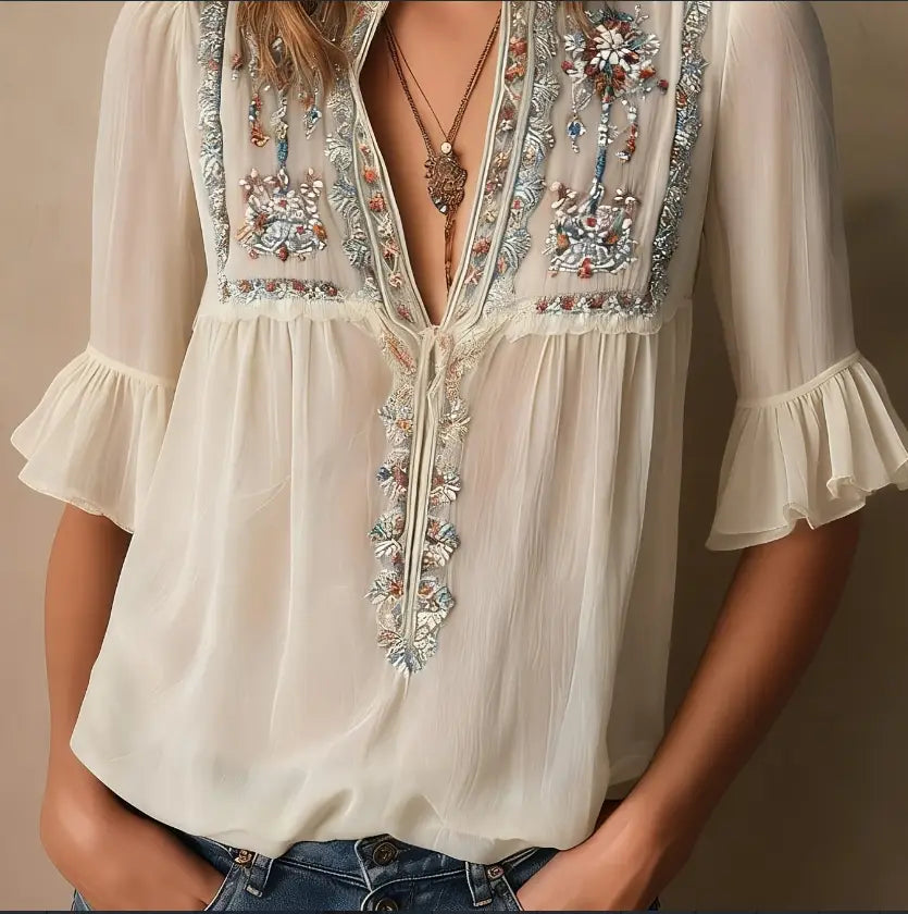 Blouse en mousseline de soie ivoire délicate, présentant une broderie florale complexe dans des tons bleu argenté et rose le long de l’encolure et de la patte de boutonnage.