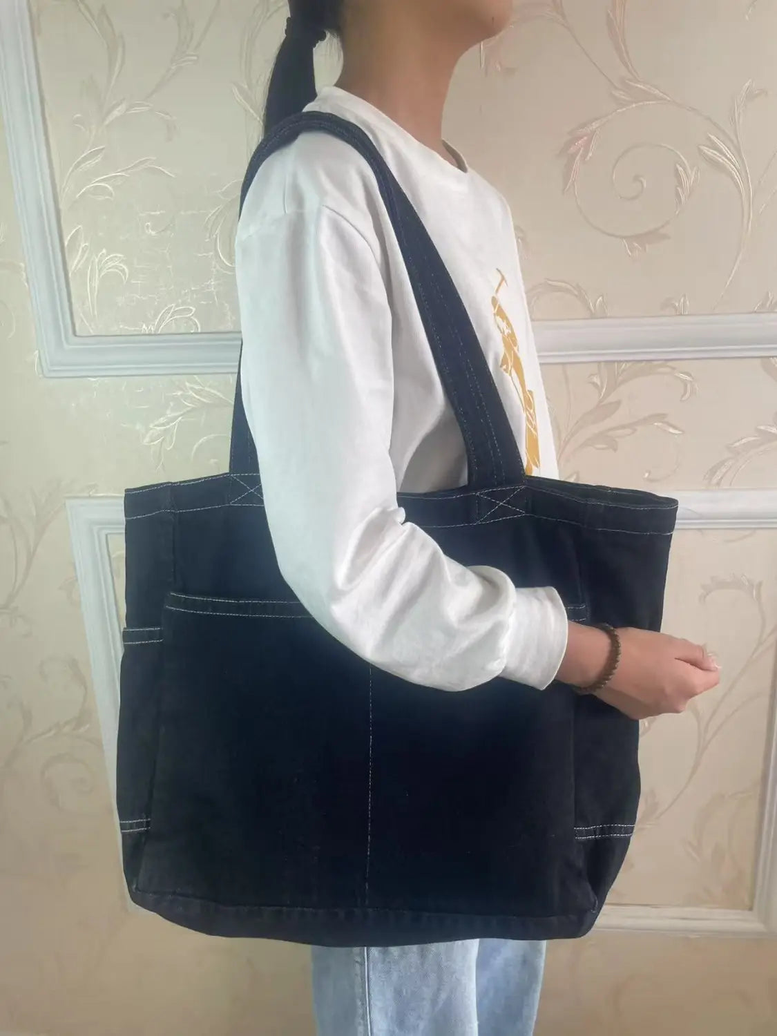 Black denim tote bag with white stitching and two front pockets designed for large capacity and shoulder use by Alma Latina.