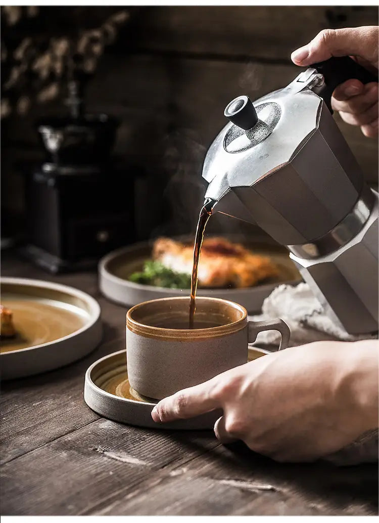 A sleek silver moka pot with a black handle pours rich coffee into a ceramic mug from a ceramic tableware set retro Japanese dinnerware.