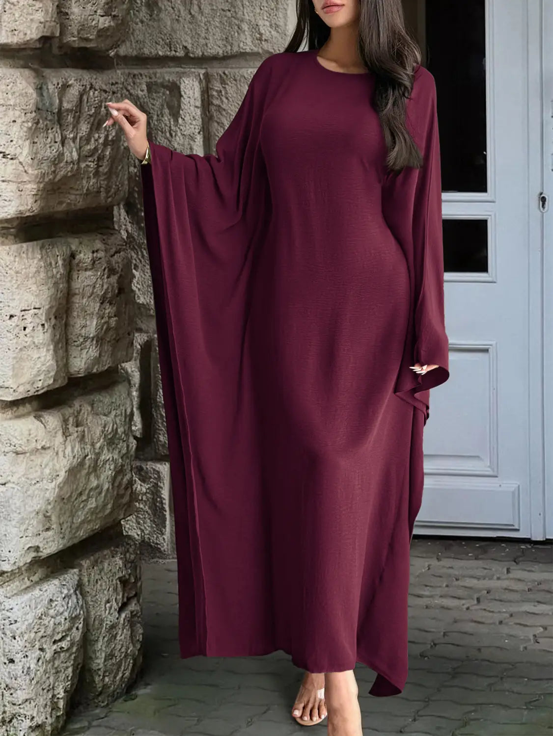 A flowing maroon satin abaya with wide loose sleeves and a round neckline styled as an elegant long sleeve satin maxi dress with ruffled hem.