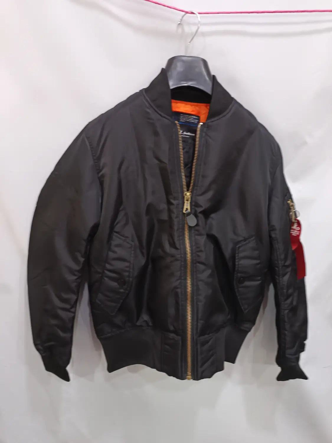 Men’s pilot cotton bomber jacket in black with a gold zipper and a red patch on the sleeve hanging on a hanger.