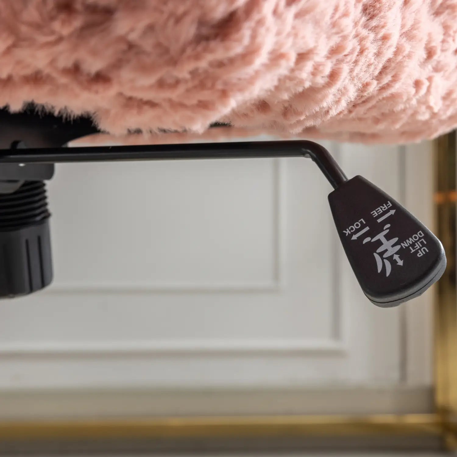 Un levier ergonomique noir de réglage de la hauteur, avec des mentions « up » (monter), « lift » (soulever), « down » (descendre) et « free lock » (libre/verrouillage) visibles sur le côté d’une chaise de bureau en fausse fourrure rose dotée d’un piétement en métal doré.