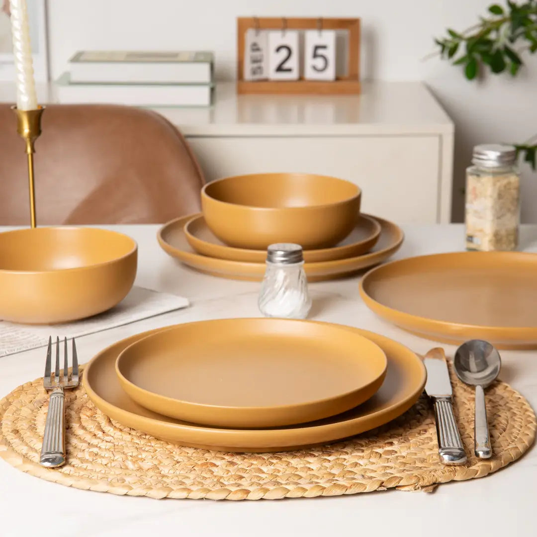 Set of matte mustard-yellow stoneware dinnerware with matching bowls and plates arranged on a woven placemat for four people.