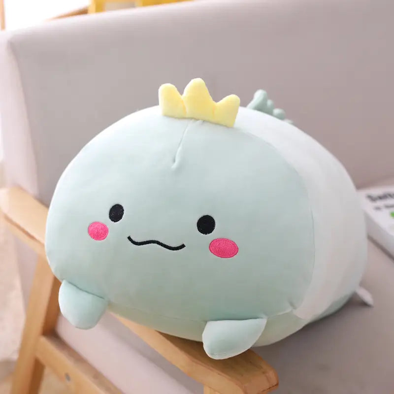 A soft light blue plush animal pillow in the shape of a dinosaur with a yellow crown and pink cheeks designed as a cartoon doll for kids.