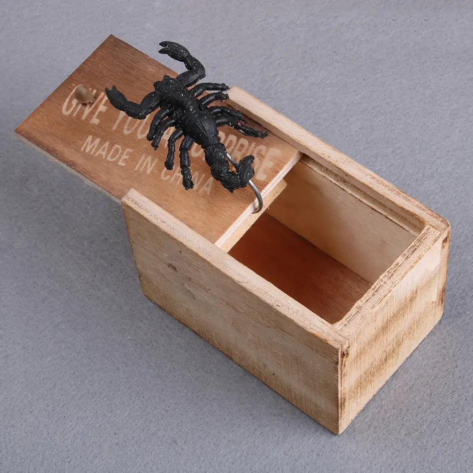 薄い茶色の木製の「いたずらの見せかけ箱」の上に止まった黒いサソリのフィギュアで、刻印された文字があしらわれ、いたずらのサプライズ玩具として設計されている。