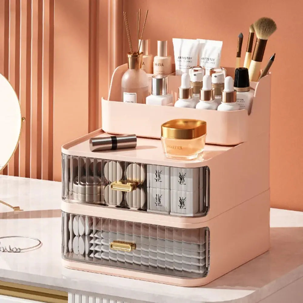 Blush pink two-tiered desktop makeup and jewelry organizer with transparent drawers and gold-tone handles holding beauty products