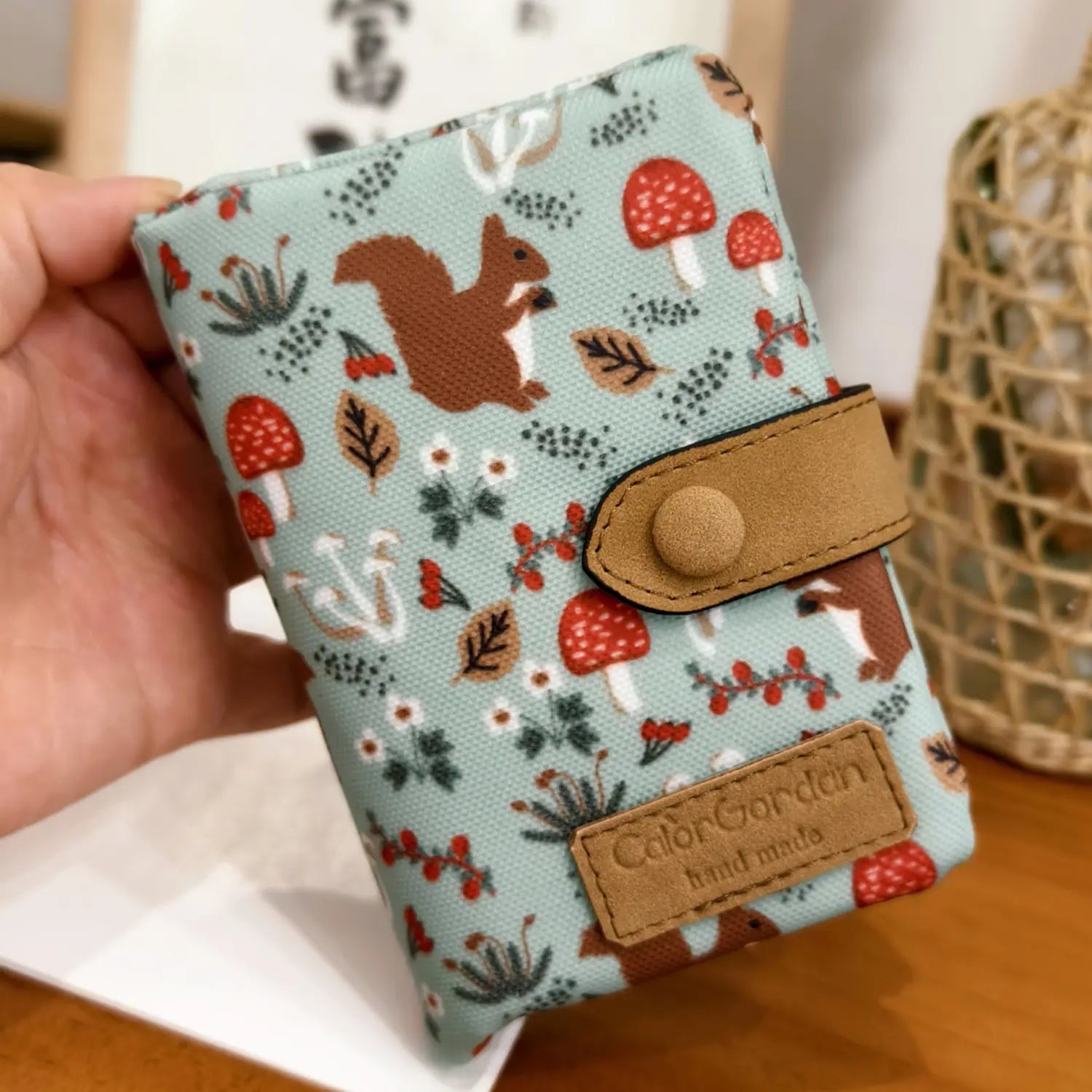 A light blue canvas tri fold multi card wallet featuring a whimsical pattern of red mushrooms brown squirrels and autumn leaves accented with tan leather straps and a matching branded patch.