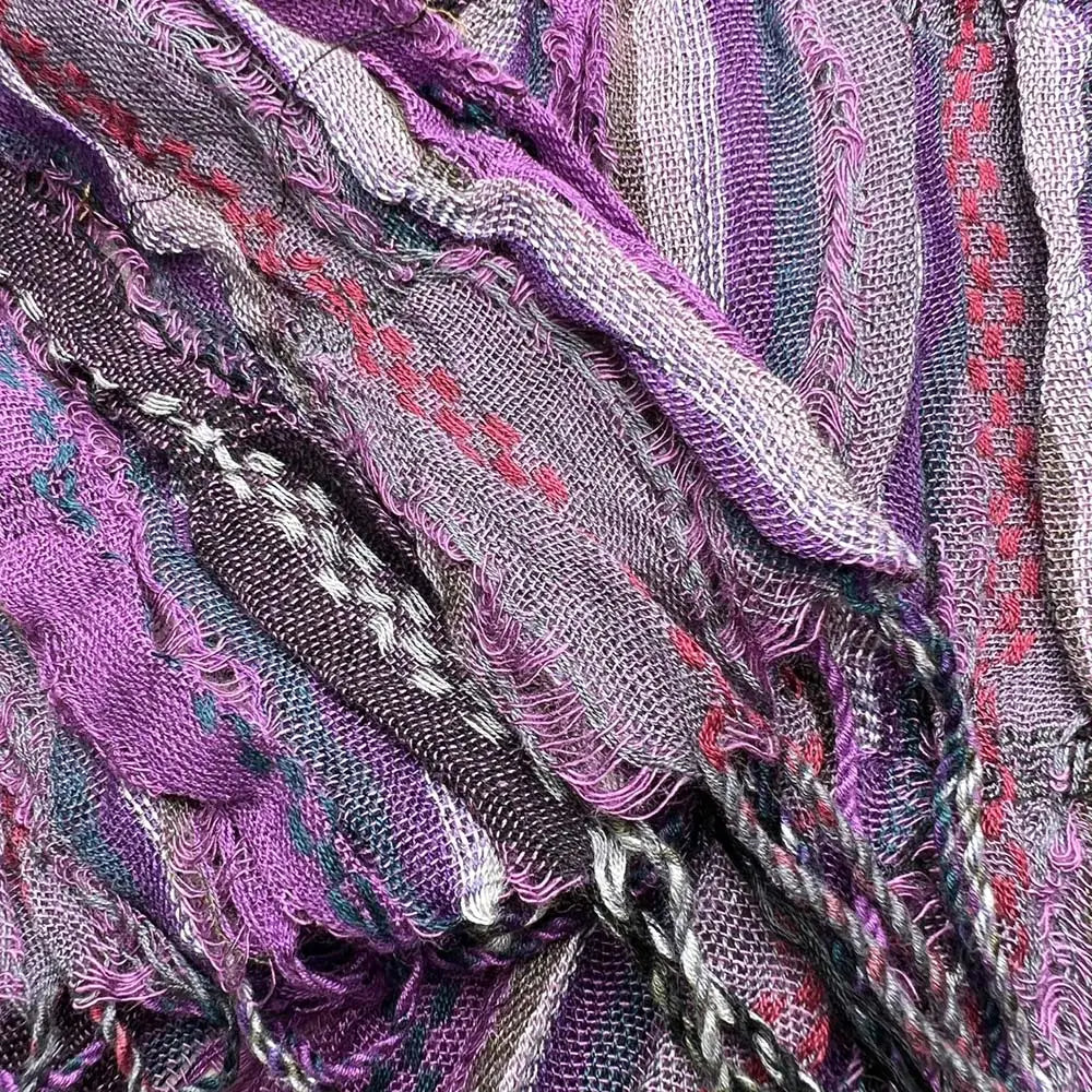 Une écharpe froissée en jacquard violet texturé, aux bords effilochés et aux motifs tissés dans des nuances de violet, de gris, de rose et de noir, conçue comme un accessoire d’hiver.