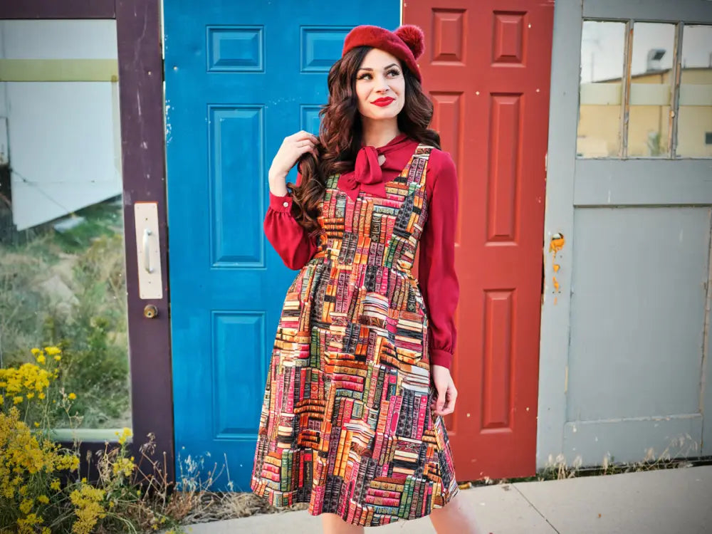 A vibrant bookshelf print cotton fit and flare dress with pockets worn over a red long-sleeved blouse and matching beret