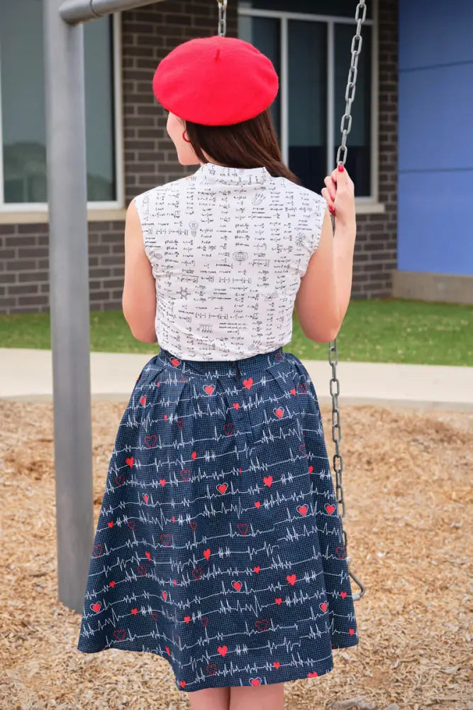 A woman wearing a navy blue Doris navy pleated heartbreaker skirt with pockets featuring red hearts and ECG line pattern