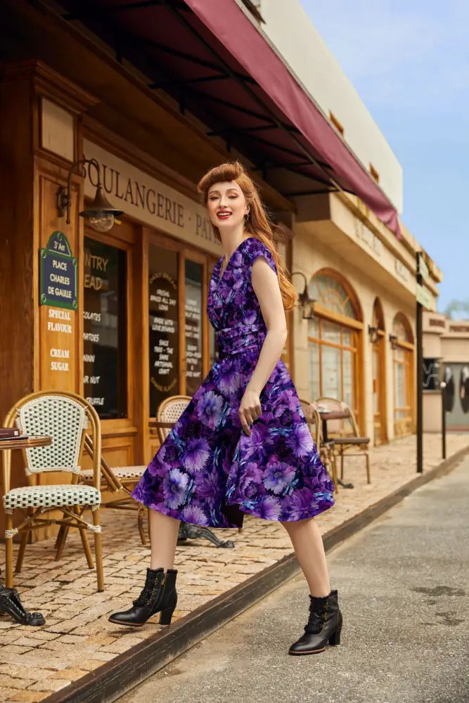 A woman wearing a vibrant purple cotton dragonfly floral greta v-neck flare dress with bold violet and magenta blossoms