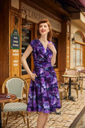 A woman wears a dragonfly floral greta v-neck cotton flare dress in vibrant purple with violet and blue flower patterns