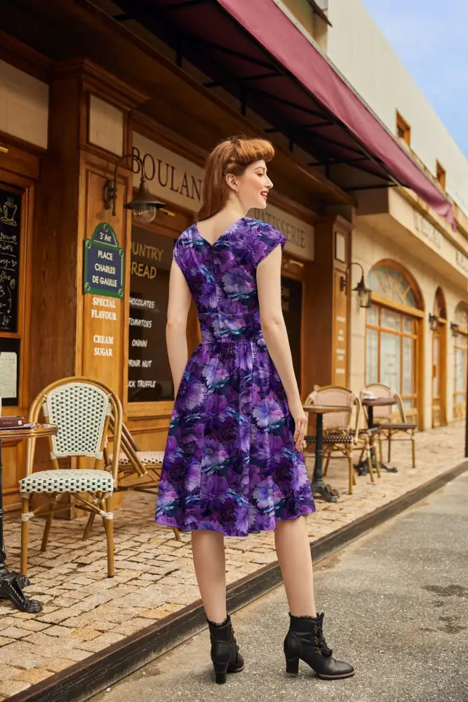 A woman wears a purple floral print dragonfly floral greta v-neck cotton flare dress with short sleeves and fitted waist