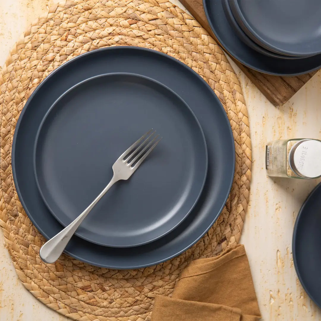 Set of navy blue ceramic matte finish plates stacked on a woven placemat from a 12-piece stoneware dinnerware set for three.