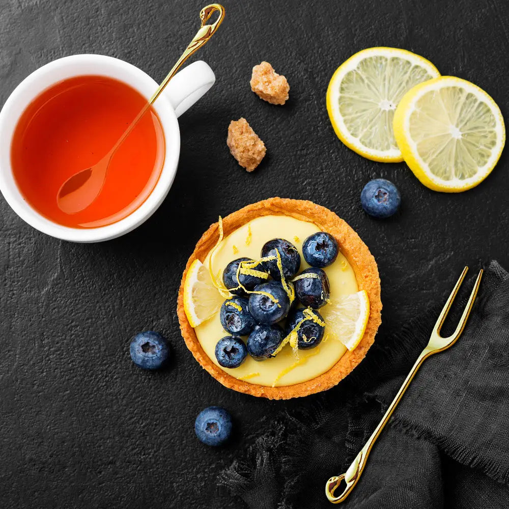 Une tartelette dorée citron-myrtille, avec une garniture de crème jaune dans une pâte croustillante, rehaussée de myrtilles fraîches et de zeste de citron, aux côtés d’un Coffret de coutellerie Cygne en cuillères et fourchettes à dessert dorées.