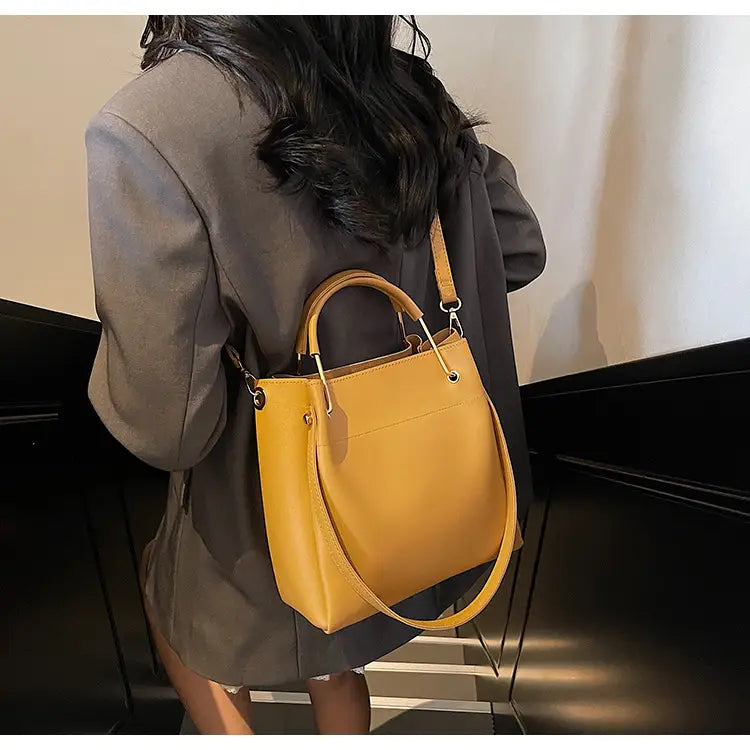 A mustard-yellow minimalist leather bucket bag with a structured shape featuring dual top handles and an adjustable detachable shoulder strap.