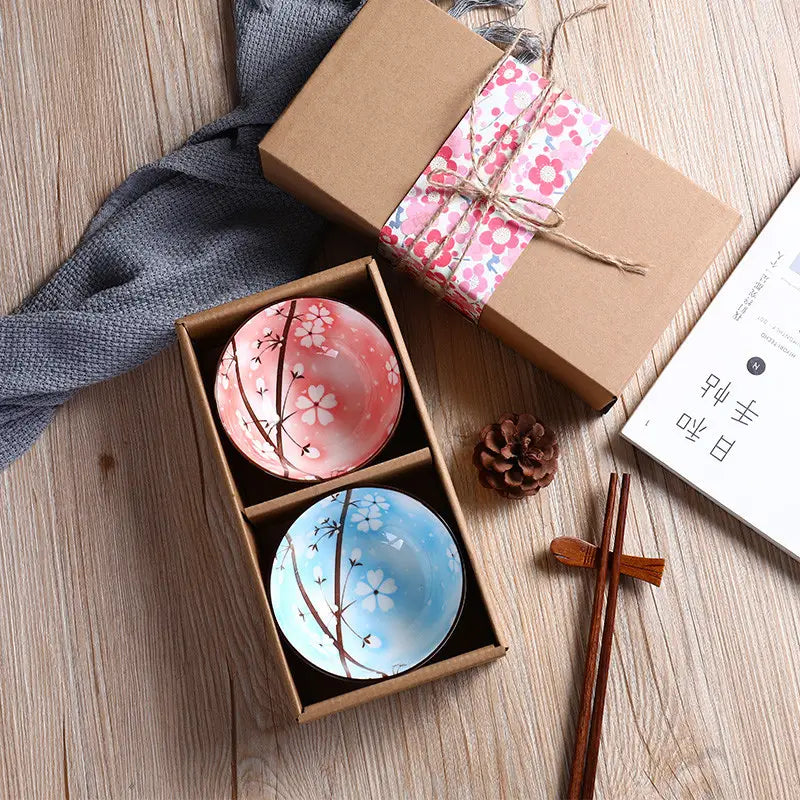 A pair of ceramic bowls from a Japanese tableware set featuring cherry blossom designs with one bowl in soft pink and the other in light blue, each with delicate dark brown branches.